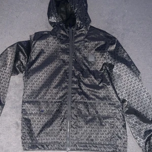 Michael kors jacka - Snygg grå vindjacka från Michael Kors med ett stilrent MK-mönster. Jackan har dragkedja framtill och en praktisk huva. Perfekt för blåsiga dagar och ger en trendig look.