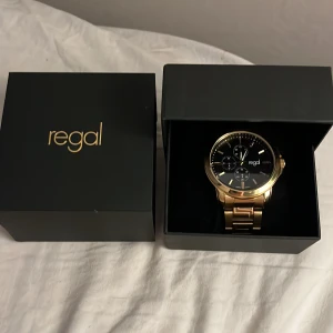 Guldfärgad klocka från Regal - Snygg guldfärgad klocka från Regal med svart urtavla och flera små urtavlor. Armbandet är i metall och matchar den eleganta designen. Perfekt för den som vill ha en stilren accessoar.