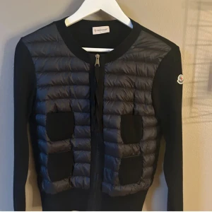 Svart jacka från Moncler - Snygg svart jacka från Moncler med quiltad design och dragkedja framtill. Jackan har långa ärmar och en stilren look med ribbade detaljer vid ärmslut och nederkant. Perfekt för en trendig och bekväm stil.