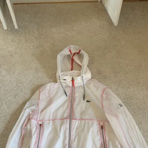 Vit vindjacka från Lacoste Sport - Säljer en stilren vit vindjacka från Lacoste Sport med röd dragkedja och justerbar huva. Jackan har det klassiska Lacoste-märket på bröstet och är en del av Novak Djokovic-kollektionen. Perfekt för sportiga aktiviteter eller en avslappnad look.