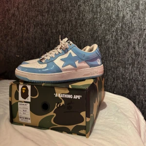 Blåa Bapesta - Snygga blå och vita sneakers från A Bathing Ape med en glansig finish och den ikoniska stjärndetaljen på sidan. Skorna har snörning och en robust sula med stjärnmönster undertill. Perfekta för att ge din outfit en unik touch. Den är dock lite cooked men fortfarande användbar. Bape påsen till dem finns inte