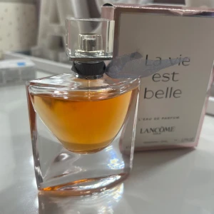 La Vie Est Belle Eau de Parfum från Lancôme - Elegant parfymflaska med en varm, bärnstensfärgad doftvätska. Flaskan har en stilren design med en liten rosett vid halsen. Förpackningen är vit med rosa detaljer och texten 'La Vie Est Belle'. Perfekt för den som älskar en sofistikerad och feminin doft.