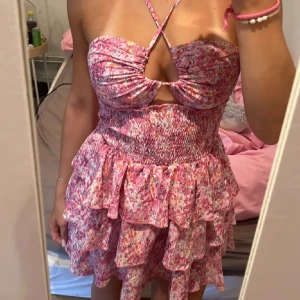 Rosa blommig klänning - Söt rosa blommig klänning med volanger och smockdetaljer. Axelbandslös design med knyt framtill och kort längd. Perfekt för sommaren!