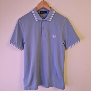 Ljusblå pikétröja från Fred Perry - Säljer en stilren ljusblå pikétröja från Fred Perry med klassisk krage och vita detaljer. Tröjan har korta ärmar och är perfekt för en avslappnad stil. Den ikoniska loggan är broderad på bröstet.