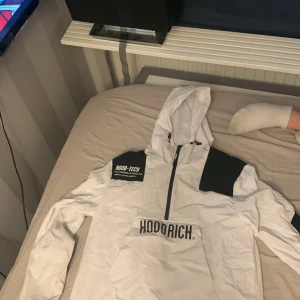Vit vindjacka från Hoodrich - Säljer en vit vindjacka från Hoodrich med svart text på bröstet och ärmen. Jackan har en justerbar huva och dragkedja framtill. Perfekt för en streetwear-look.