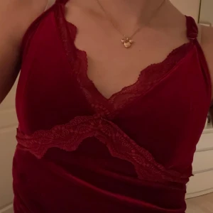 Röd klänning med spetsdetaljer - Snygg röd klänning med spetsdetaljer och korsade axelband. Klänningen har en omlott-design med asymmetrisk nederkant och är tillverkad i ett glansigt material som ger en lyxig känsla.