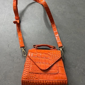 Orange axelväska med krokodilmönster - Snygg orange axelväska med krokodilmönster och justerbar axelrem. Väskan har ett handtag och en praktisk ficka framtill. Perfekt för att ge en färgklick till din outfit!