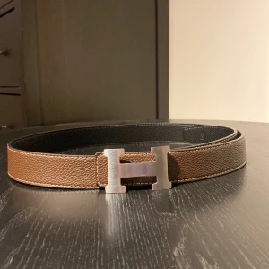 Brunt och svart bälte från Hermès - Snyggt vändbart bälte från Hermès i brunt och svart skinn med en ikonisk H-formad spänne i silver. Perfekt för att ge en lyxig touch till din outfit. Bältet har en elegant och tidlös design.