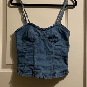 Blå jeanskorsett med volangdetaljer!  - Snygg blå jeanskorsett med justerbara axelband och volangdetaljer längs kanterna. Korsetten har en dragkedja bak och är perfekt för en trendig look. Passar bra till både kjol och byxor.  Ljust foder inuti.  Översta öglan har lossnat ( medföljer vid köp)  Men plagget fungerar fint utan den!  Otroligt coolt snyggt och fint plagg!