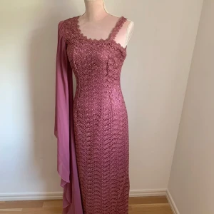 Rosa spetsklänning med cape - Elegant rosa långklänning i spets med en unik oneshoulder-design och en dramatisk cape. Perfekt för speciella tillfällen där du vill sticka ut. Klänningen har en figurnära passform som framhäver silhuetten.