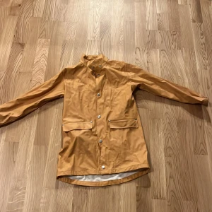 Orange/gul regnjacka från lager 157 i storlek 130-140 - Snygg orange/gul regnjacka från lager 157 med tryckknappar och två praktiska fickor framtill. Jackan har en stilren design och är perfekt för regniga dagar. Den har långa ärmar och en bekväm passform.