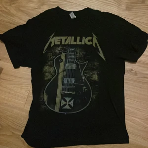 Svart Metallica t-shirt med gitarrmotiv - Cool svart t-shirt med Metallica-logga och ett stort gitarrmotiv på framsidan. Perfekt för rockälskare och fans av bandet. Kortärmad och i en klassisk passform. Inga defekter
