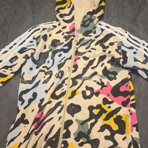 Camouflage hoodie från Adidas - Säljer en cool hoodie från Adidas med ett unikt camouflage-mönster i beige, rosa, gul och svart. Den har dragkedja och huva, samt de klassiska tre ränderna på ärmarna. Perfekt för en sportig look! Ny har kanske används en gång!