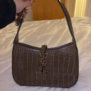 Brun axelväska med krokodilmönster - Snygg brun axelväska med krokodilmönster och elegant YSL i guld på framsidan. Den kommer inte till användning och har inte gjort det sedan jag köpte den så därför säljer jag den.
