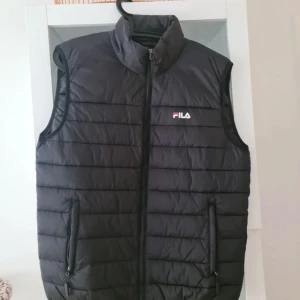 Svart dunväst från FILA - Snygg svart dunväst från FILA med dragkedja framtill. Västen har en hög krage och två fickor med dragkedja. Perfekt för kyligare dagar.