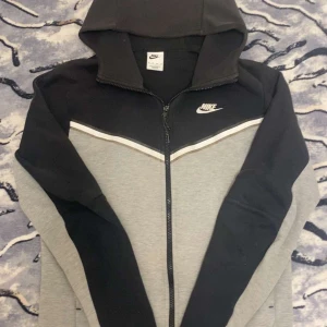 Grå och svart hoodie från Nike - Säljer en snygg grå och svart hoodie från Nike med dragkedja och huva. Den har en vit randdetalj över bröstet och Nike-loggan på vänster sida. Perfekt för en sportig look!