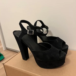 Svarta högklackade sandaler från Nelly - Snygga svarta sandaler med hög klack och öppen tå. De har en justerbar rem med spänne runt ankeln för en säker passform. Perfekta för en kväll ute eller en speciell tillställning.