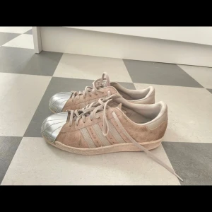 Beige Adidas Superstar med silverdetaljer - Snygga beige Adidas Superstar sneakers med silverfärgad tåhätta och klassiska ränder. Skorna har snörning och är tillverkade i mocka med en stilren design.