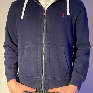 Mörkblå ziphoodie från Polo Ralph Lauren - Snygg mörkblå hoodie från Polo Ralph Lauren med dragkedja och vit snörning i huvan. Den har en klassisk röd logga på bröstet och praktiska fickor framtill. Perfekt för en avslappnad stil.
