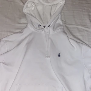 Vit hoodie från Polo Ralph Lauren - Säljer en stilren vit hoodie från Polo Ralph Lauren med det klassiska logotypbroderiet på bröstet. Perfekt för en avslappnad look. Den har en justerbar huva med dragsko och en stor ficka framtill.