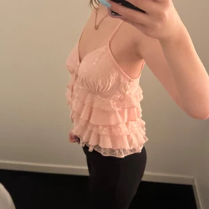 Rosa volangtopp med spetsdetaljer - Säljer en söt rosa topp med volanger och spetsdetaljer. Toppen har smala axelband och en v-ringad design. Perfekt för en somrig look!