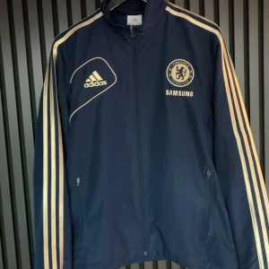 Chelsea fc Träningsjacka - Snygg mörkblå  träningsjacka med guldiga ränder från Adidas med Chelsea FC-logga. Jackan har långa ärmar och dragkedja framtill. Den är från 12/13 säsongen.