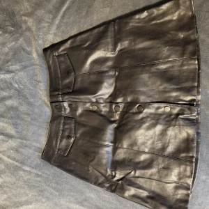 Svart skinnkjol från Divided - Snygg svart skinnkjol från Divided med knappar framtill. Kjolen har en klassisk A-linjeform och är perfekt för en trendig look. Den är tillverkad i ett glansigt material som ger en cool och stilren känsla.