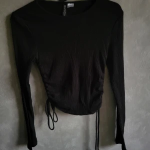 Svart topp från H&M Divided - Säljer en svart topp från H&M Divided med långa ärmar och knytband i midjan. Perfekt för en stilren look. Passar bra till både jeans och kjol.