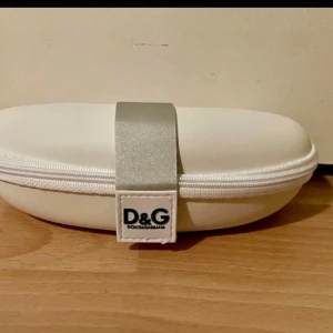 Vit glasögonfodral från Dolce & Gabbana - Elegant och stilrent vitt glasögonfodral från Dolce & Gabbana. Fodralet har en mjuk insida och en dragkedja för säker förvaring. Perfekt för att skydda dina glasögon med stil. 