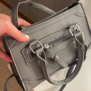 Grå axelväska med dragkedja - Snygg grå axelväska i syntetmaterial med praktisk dragkedja. Perfekt för att bära dina dagliga nödvändigheter. Stilren design med dubbla handtag och justerbar axelrem.