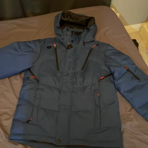 Blå dunjacka med huva - Säljer en blå dunjacka med huva och flera dragkedjefickor. Jackan har en stilren design med svarta detaljer och är perfekt för kyligare väder. Har ett litet klipp håll i naken kan sys mycket lätt många kommer skriva passa på 