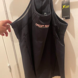 Svart linnetopp från Drop Shot - Säljer en svart linnetopp från Drop Shot, perfekt för träning. Toppen har en sportig design med brottarrygg och är tillverkad i ett lätt och andningsbart material.