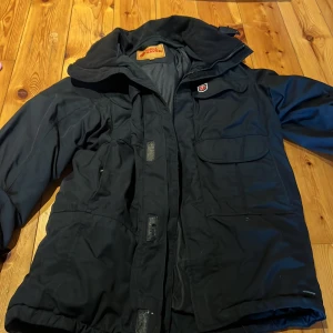 Svart jacka från Fjällräven - Säljer en svart Fjällräven jacka med dragkedja och knappar. Jackan har en praktisk design med flera fickor och en broderad logotyp på bröstet. Perfekt för kyligare väder.