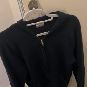 Mörkblå kashmir hoodie från John Henric - Säljer en stilren mörkblå hoodie i mjuk kashmir från John Henric. Tröjan har en dragkedja framtill och en bekväm huva. Perfekt för kyliga dagar. Litet hål vid baksidan (kolla bilden), inte jätteviktigt för ingen använder hoodien med luva ändå. Skick 9/10