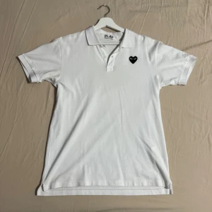 Vit pikétröja från Comme des Garçons - Säljer en vit pikétröja från Comme des Garçons med det ikoniska hjärtemärket på bröstet. Tröjan har korta ärmar och en klassisk krage med knappar. Perfekt för en minimalistisk look.