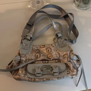 Beige och grå axelväska med mönster - As snygg axelväska från Gussaci i beige och grått med logotypmönster. Väskan har flera detaljer som dragkedja och spännen i silverfärg. Perfekt för att bära dina dagliga nödvändigheter med stil. köpt på semester från lång tid tillbaka🫶 Helt oanvänd