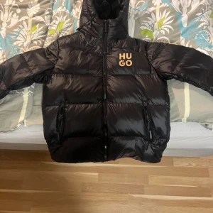 Svart pufferjacka från Hugo - Snygg svart pufferjacka från Hugo med orange logotyp på bröstet. Jackan har en dragkedja framtill och två fickor med dragkedjor. Perfekt för kyliga dagar med sin vadderade design och huva. Storlek S . 