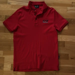 Röd pikétröja från Emporio Armani - Snygg röd pikétröja från Emporio Armani med EA7-logga på bröstet. Aldrig använd tröjan! Tröjan har en klassisk krage och knappar framtill. Perfekt för en stilren look. Hör av dig!