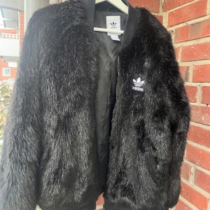 Bombarjacka i fuskpäls från adidas  - Snygg svart pälsjacka från Adidas med deras ikoniska logga på bröstet. Jackan har en mjuk och fluffig yta som ger en lyxig känsla. Perfekt för att hålla sig varm och stilren under kyligare dagar. Nypris var 3499:- 