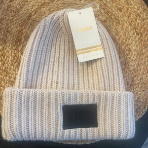 Beige stickad mössa från Stronger - Snygg beige stickad mössa från Stronger med en bred uppvikt kant och en svart etikett framtill. Perfekt för kyliga dagar och ger en stilren look.