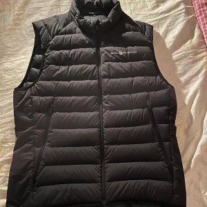 Svart dunväst från Sail Racing - Säljer en svart dunväst från Sail Racing i storlek medium. Västen har en dragkedja framtill och är quiltad för extra värme. Perfekt för kyligare dagar.