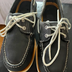 Brand: TimberlandTimberland Men's Cedar Bay 2 Eye BoatFashion Boot - Snygga svarta seglarskor med vit snörning och kontrasterande sömmar. Skorna har en klassisk design med en vit sula som ger en stilren look. Perfekta för en avslappnad stil.