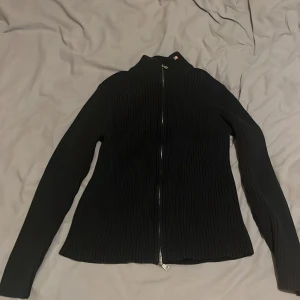 Svart ribbad RL full zip - Snygg svart ribbad tröja från RL  med dragkedja framtill. Tröjan har en hög krage och långa ärmar, perfekt för kyligare dagar. Den har en liten flaggdetalj vid kragen som ger en extra touch. Size L men passar S
