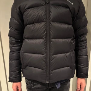 Svart dunjacka från Sail Racing storlek 150 - Snygg svart dunjacka från Sail Racing med dragkedja och huva. Perfekt för kyliga dagar med sin vadderade design. Jackan har en stilren look och är idealisk för både stad och natur. Liten lagning ( se bild) , knappt synlig och påverkar ej funktionen. 