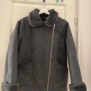 Grå jacka med fuskpäls - Snygg grå jacka med mjuk fuskpäls på insidan och i fickan. Jackan har en asymmetrisk dragkedja och är perfekt för kyliga dagar. Den har en stilren design med en bekväm passform. Har haft på den bara några gånger! 