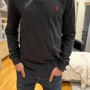 Svart långärmad pikétröja från Ralph Lauren - Säljer en stilren svart långärmad pikétröja från Ralph Lauren med det klassiska röda logotypbroderiet på bröstet. Tröjan är i bomull och har en bekväm passform. Perfekt för en avslappnad men ändå snygg look.
