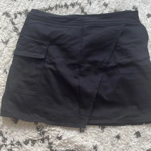 Svart kjol från Lindex - Säljer en svart kjol från Lindex i storlek 38 (M). Kjolen har en omlott-design och en praktisk ficka på sidan. Perfekt för både vardag och fest! 🖤