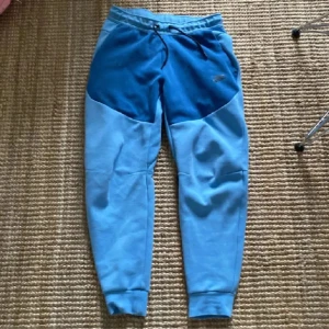 Blå mjukisbyxor från Nike - Säljer ett par blå mjukisbyxor från Nike i storlek S. Byxorna har en snygg tvåfärgad design med mörkare blå partier på framsidan och en dragkedja på baksidan. De har en elastisk midja med snörning för justerbar passform och är gjorda av mjukt och bekvämt material. Perfekta för träning eller en avslappnad dag hemma!