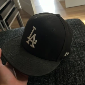 Svart keps från New Era - Säljer en svart keps från New Era med vit broderad logga på framsidan. Kepsen är i modellen 59FIFTY och har en platt skärm. Den är i bra skick och passar perfekt för både vardag och sport.