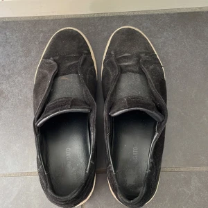 Svarta sneakers från Ahigato - Säljer ett par svarta sneakers från Arigato. De är gjorda i mocka och har en vit sula. Skorna är i välanvänt skick men fortfarande bekväma och snygga. Perfekta för vardagsbruk!